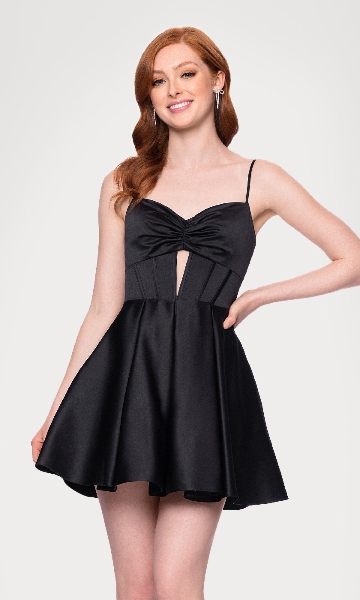 Woman with long red hair wears the Cut-Out Black Mini Hoco Dress (Blondie Nites 5605BN), a sleeveless black homecoming dress with thin straps and front knot detail, against a plain light background.